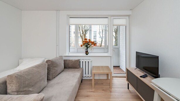A modern, minimalist living room with a sofa, a small table, a TV, and a window with a view of buildings. - Studio Ap Flora by Polo Apartments (Kaunas)