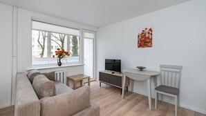 A modern living room with a sofa, TV, small table, and a window with a view of buildings. - Studio Ap Flora by Polo Apartments (Kaunas)