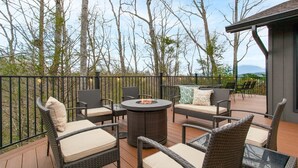 Eine Terrasse im Freien mit Korbmöbeln, einem runden Tisch und einem Feuerkorb, mit Blick auf Bäume und Berge.