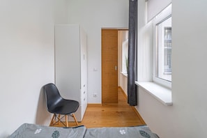 Una habitación estrecha con una cama, una silla, un armario, una ventana y una puerta que da a otra habitación.