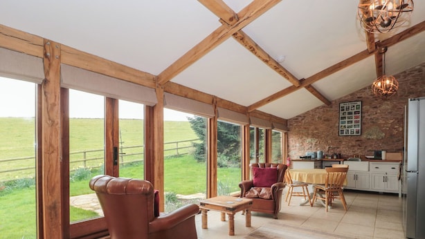 A spacious room with large windows, a wooden ceiling, a leather armchair, a dining table, and a kitchen area. - The Barn (Bridgwater)