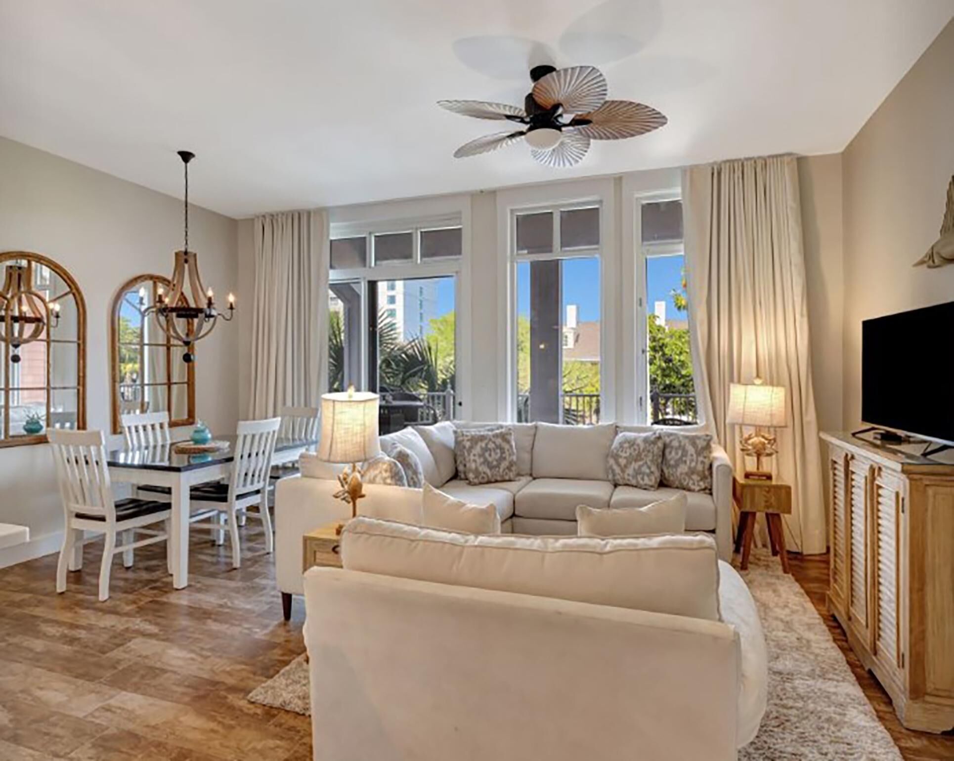 A spacious living area with a large sofa, dining table, and chairs. There is a ceiling fan, large windows with curtains, and a TV on a cabinet.
