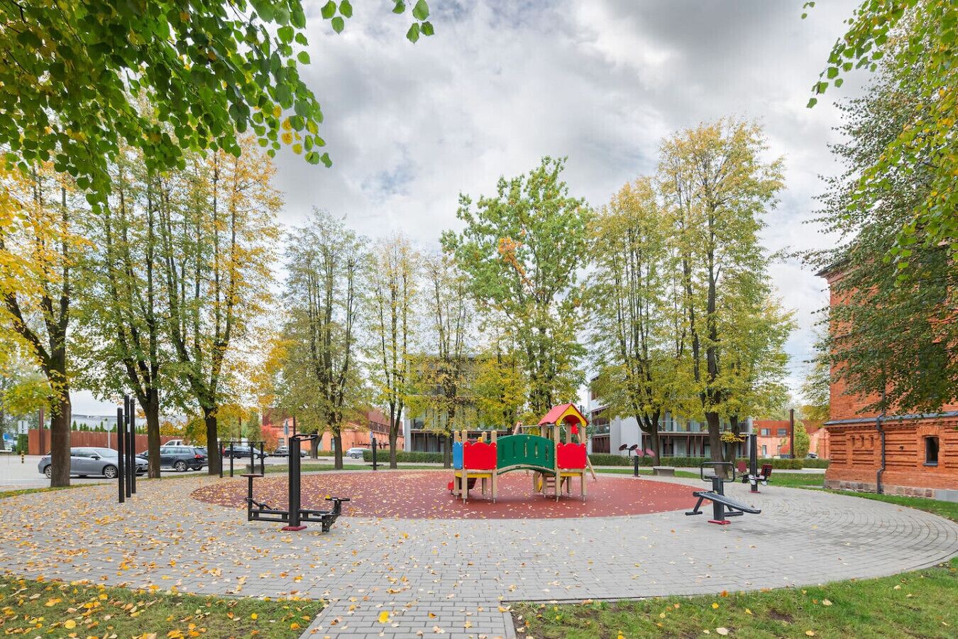 Ein Spielplatz mit einer roten und grünen Spielgeräte-Kombination, umgeben von Bäumen und einer befestigten Fläche mit herabgefallenen Blättern.