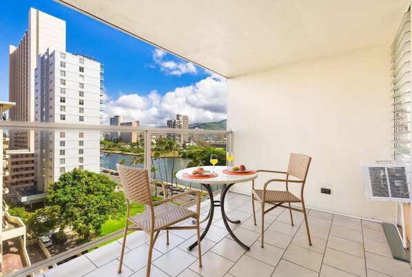 Un balcon avec deux fauteuils en osier et une petite table, d’où l’on peut admirer un paysage urbain composé d’immeubles modernes et d’une étendue d’eau.