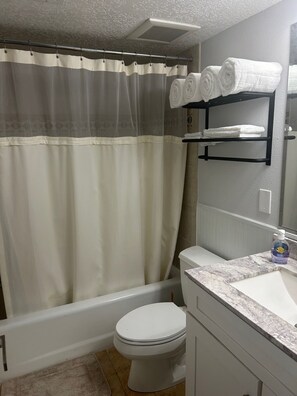 Une salle de bain avec un rideau de douche, des toilettes, un lavabo avec un plan de toilette en marbre et une étagère avec des serviettes.