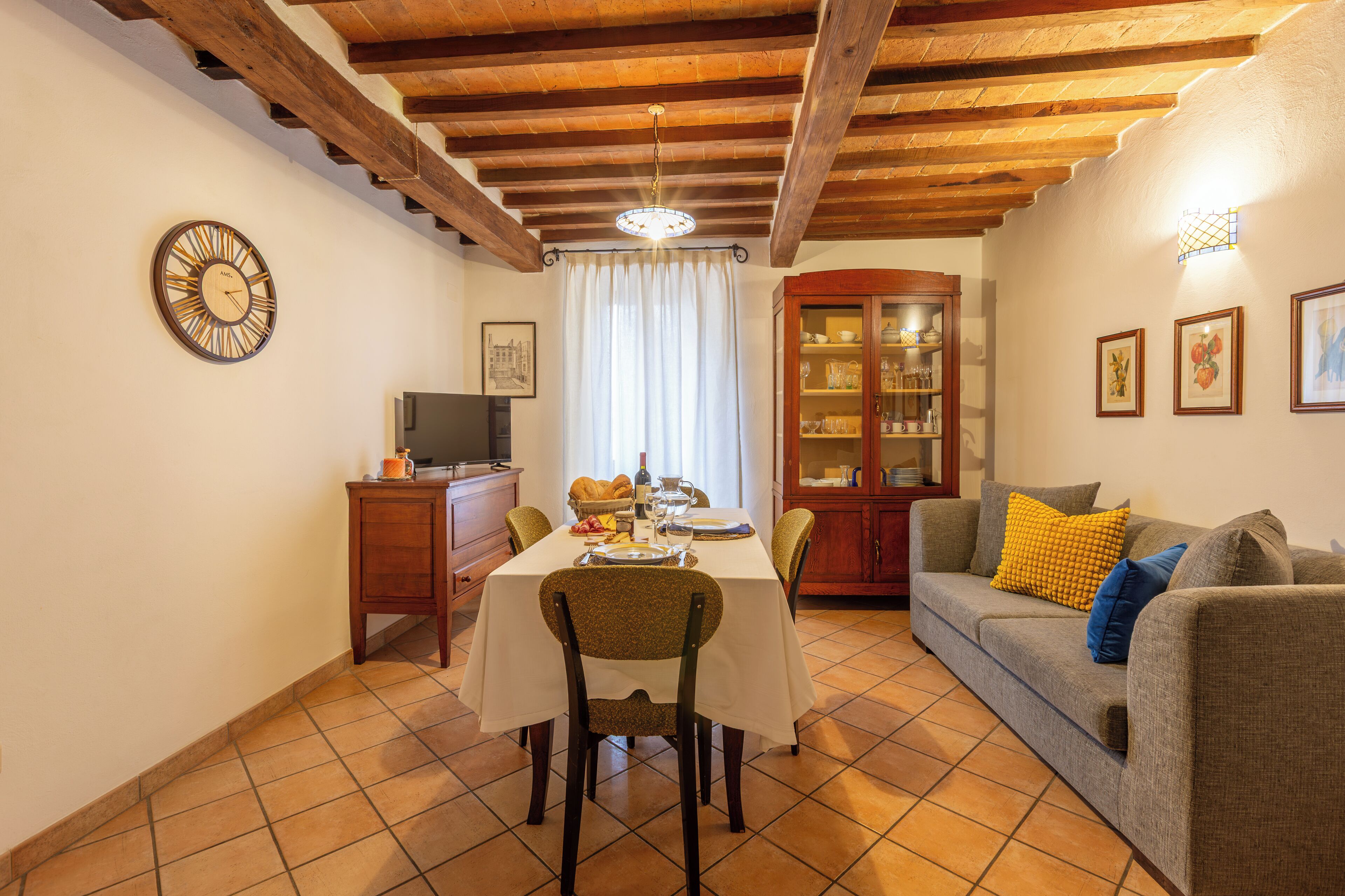 Un coin repas avec une table dressée pour deux personnes, un canapé, un meuble et une horloge au mur.