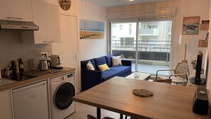 Una cocina compacta con lavavajillas Bosch, cafetera y lavadora. En la zona de estar hay un sofá azul y una mesa de comedor con sillas, junto a un amplio ventanal con vistas a la ciudad.