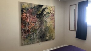 A bedroom with a large abstract painting on the wall, a bed with a purple bedspread, a ceiling fan, and a window with blue curtains. - Downtown walkable to restaurants, bars public transportation and the waterfront. (Charlottetown)