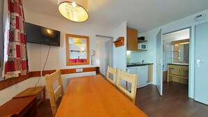 A dining area with a wooden table and chairs, a television mounted on the wall, and a kitchen in the background.