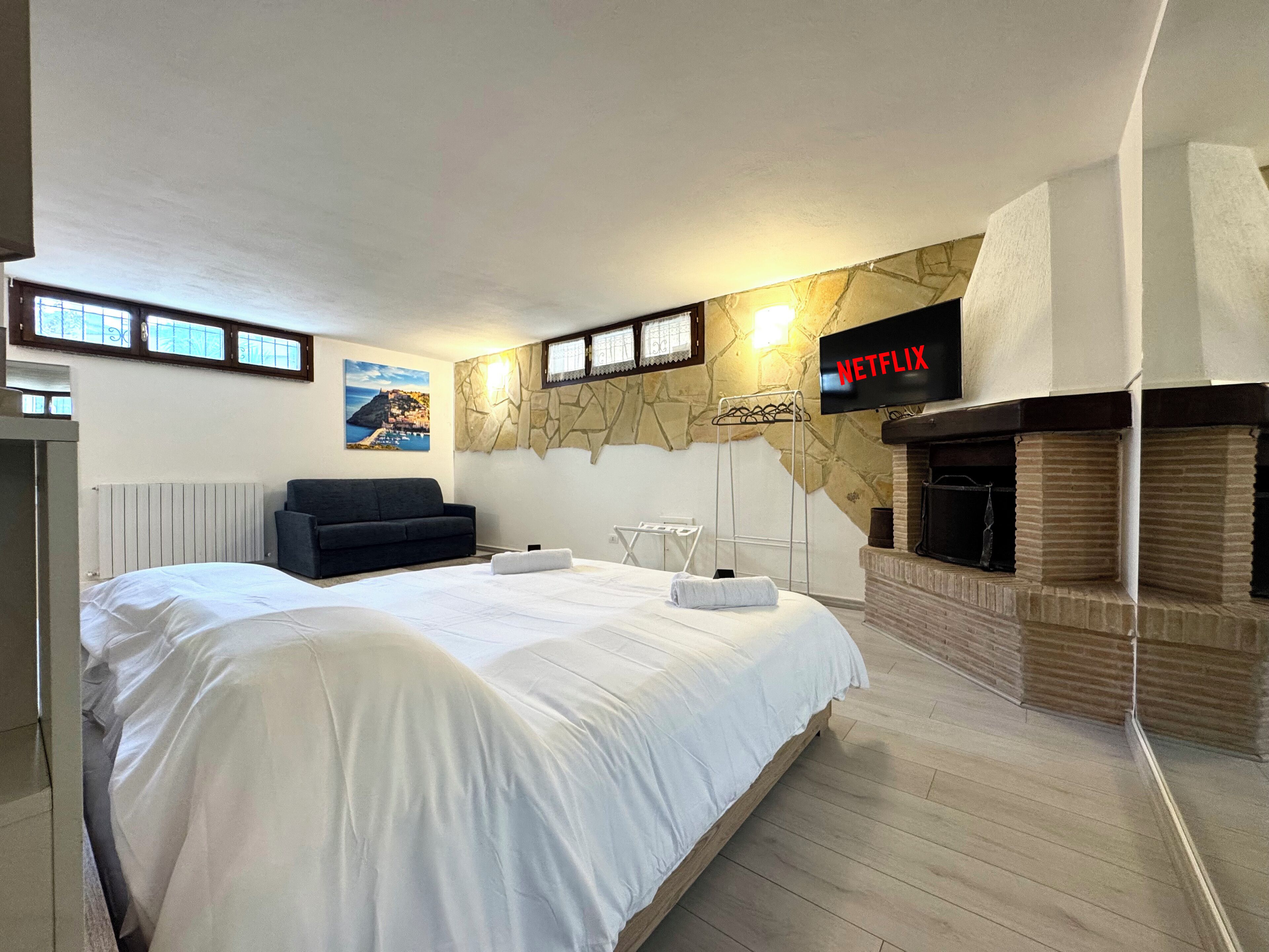A bedroom with a large bed, a black sofa, a stone wall, a fireplace, and a TV displaying 'NETFLIX'.