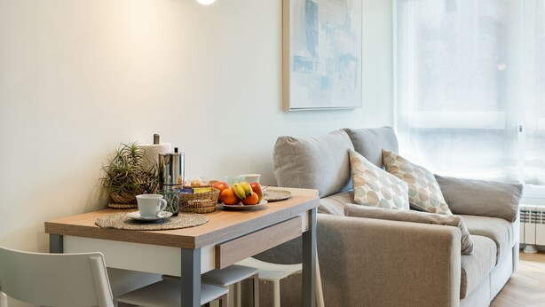 Una sala de estar moderna con comedor. Incluye un sofá, una mesa con frutas y una taza, y una lámpara colgante.