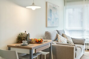 Una sala de estar moderna con comedor. Incluye un sofá, una mesa con frutas y una taza, y una lámpara colgante.