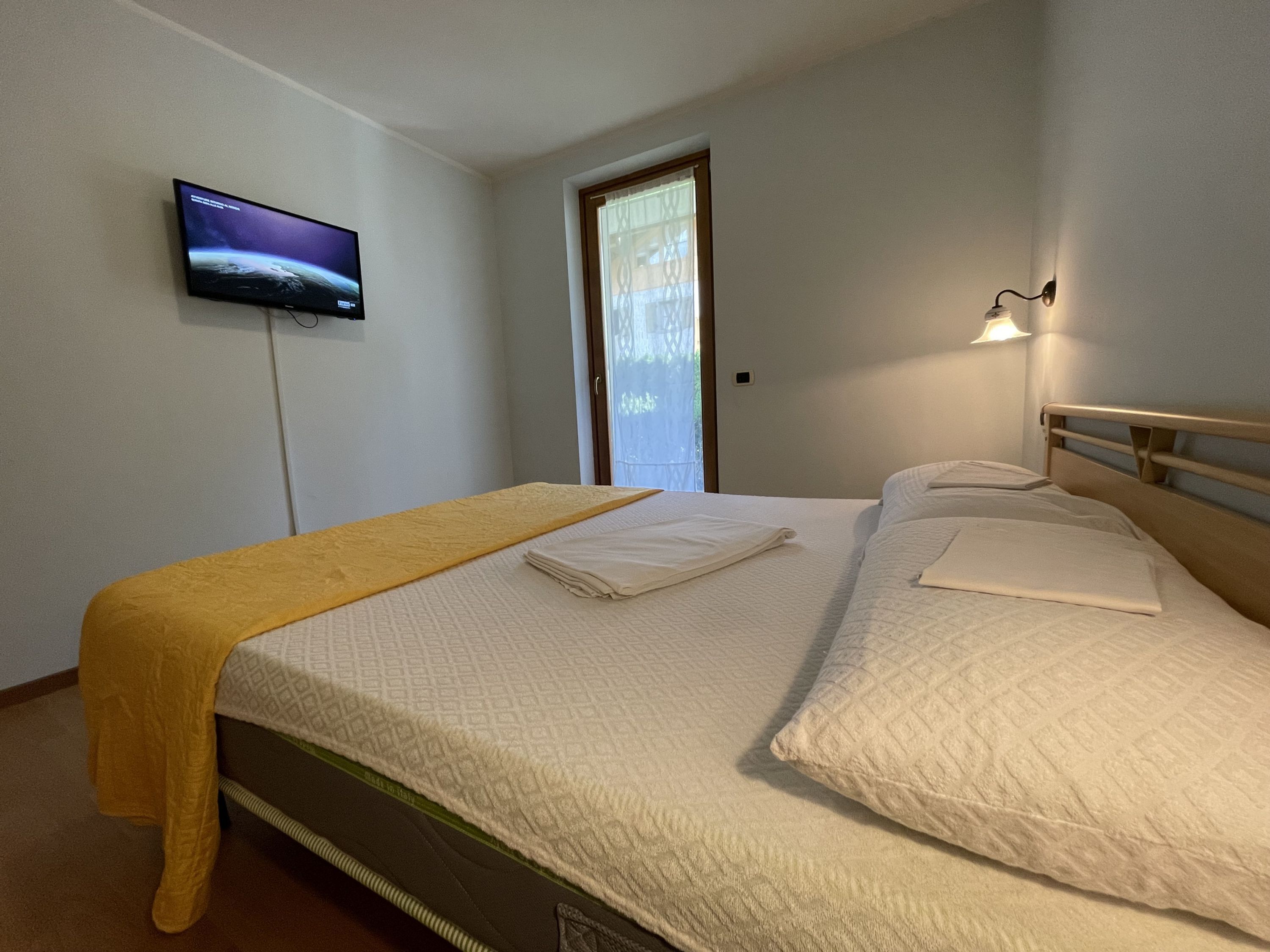 A neatly made bed with a yellow bedspread, a television mounted on the wall, and a window with curtains.