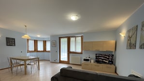 Ett rymligt vardagsrum med en soffa, ett matbord och stolar. Det finns ett köksutrymme med skåp och en mikrovågsugn. Rummet har stora fönster med gardiner och en taklampa.