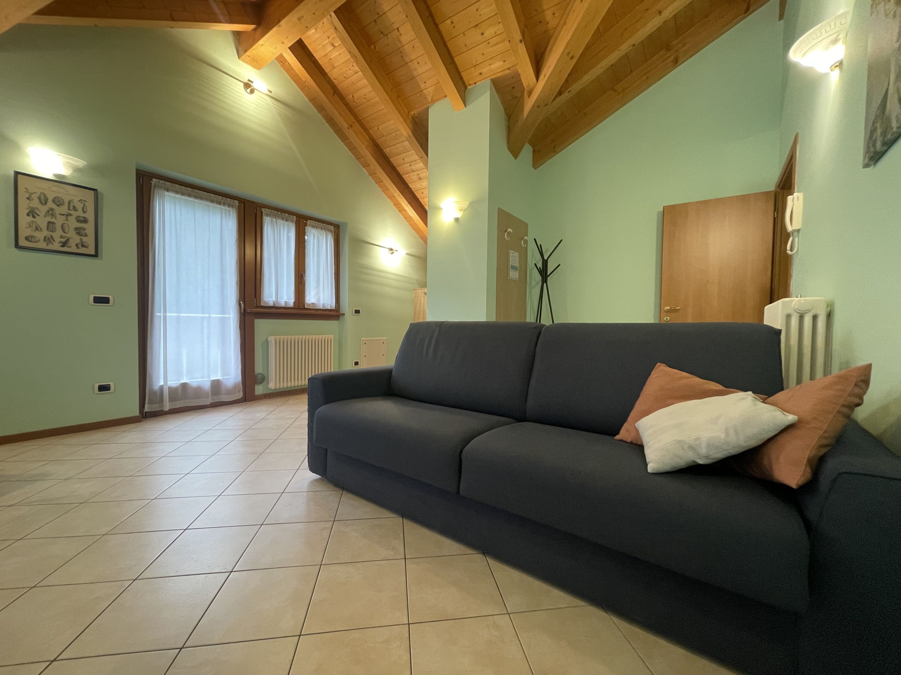 A living room with a dark sofa, a radiator, a wooden ceiling, and a window with curtains.