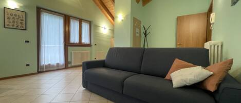 A living room with a dark sofa, a radiator, a wooden ceiling, and a window with curtains.