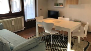 Habitación moderna con una mesa blanca, cuatro sillas, televisor, radiador y ventana con cortinas.