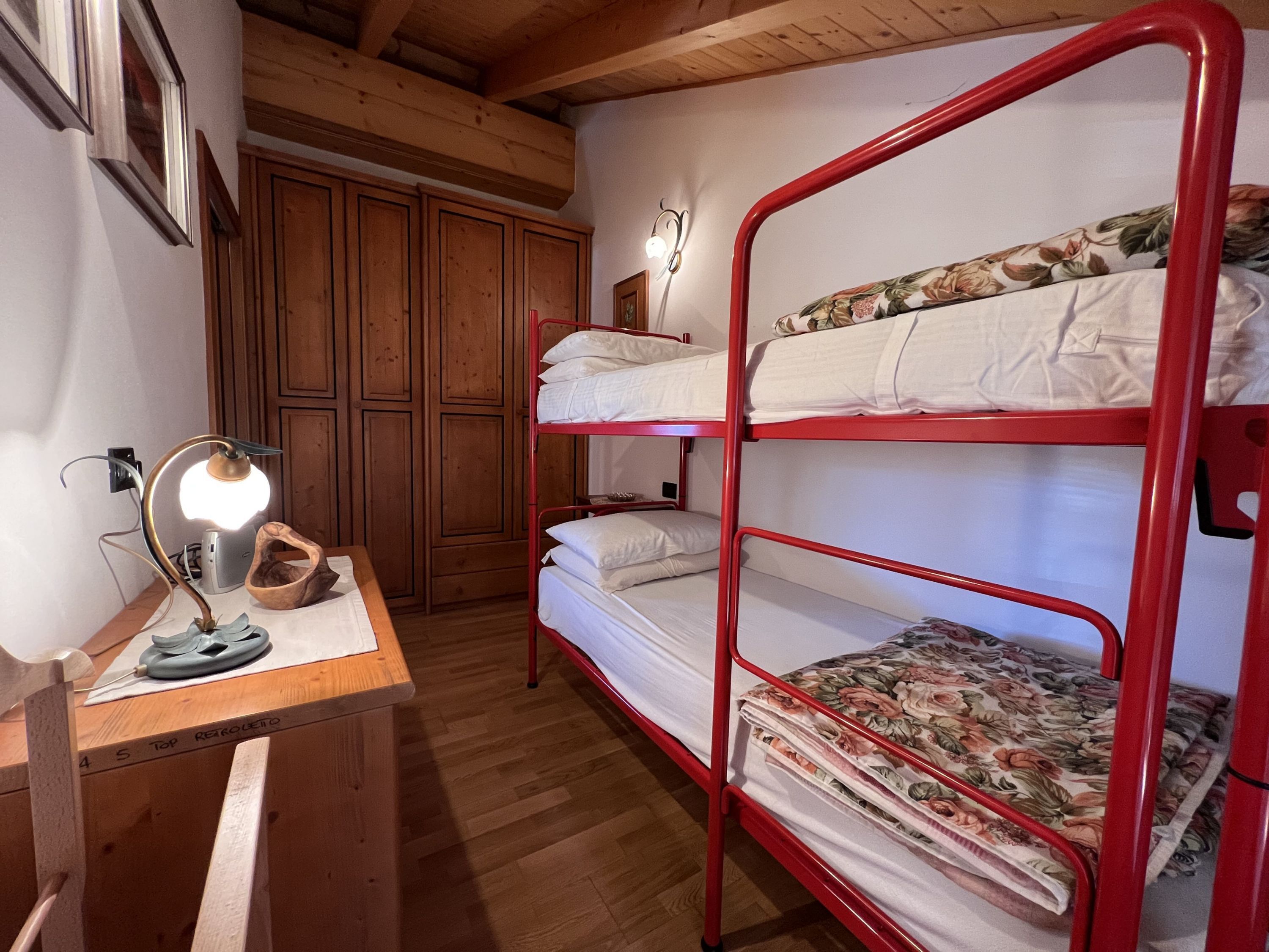 A bunk bed room with wooden flooring, a desk with a lamp, and a wardrobe.