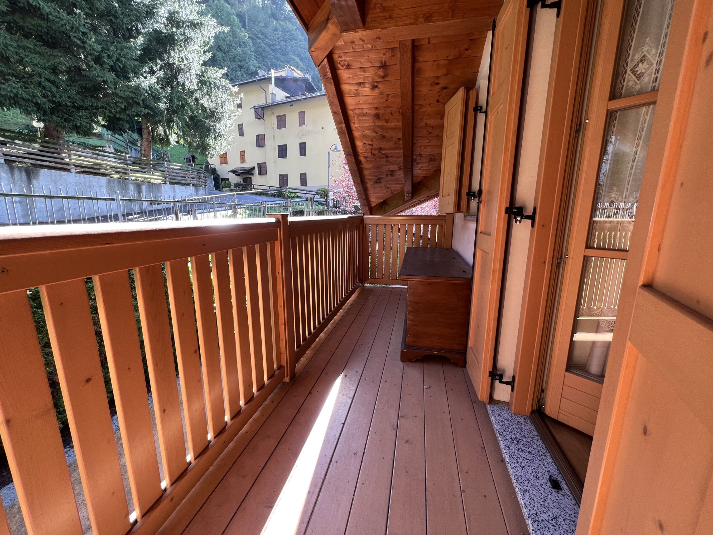 Eine Holzdielen-Terrasse mit Geländer, einer kleinen Sitzecke und Blick auf ein Gebäude sowie Bäume im Hintergrund.