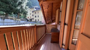 A wooden deck with a railing, a small seating area, and a view of a building and trees in the background. - 004 Trilocale di Charme Giustino (Giustino)