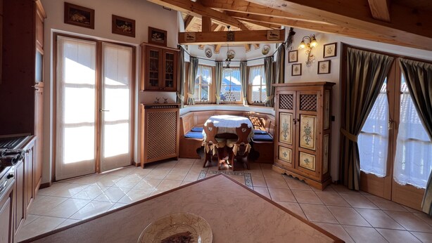 A cozy room with wooden beams, a dining table with chairs, a kitchen area, and a large window with curtains. - 004 Trilocale di Charme Giustino (Giustino)