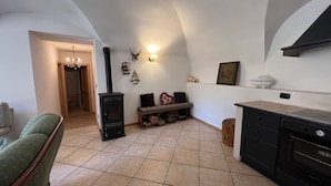 A cozy living room with a fireplace, a sofa, a bench, and a kitchen area with a stove and cabinets. - 007 Trilocale con Giardino Giustino (Giustino)