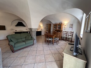 A living room with a green sofa, wooden dining table, and chairs, a cabinet, and a television. - 007 Trilocale con Giardino Giustino (Giustino)