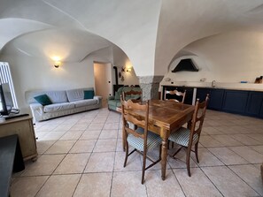 A spacious living area with a wooden dining table, a sofa, and a kitchen with a built-in oven. - 007 Trilocale con Giardino Giustino (Giustino)