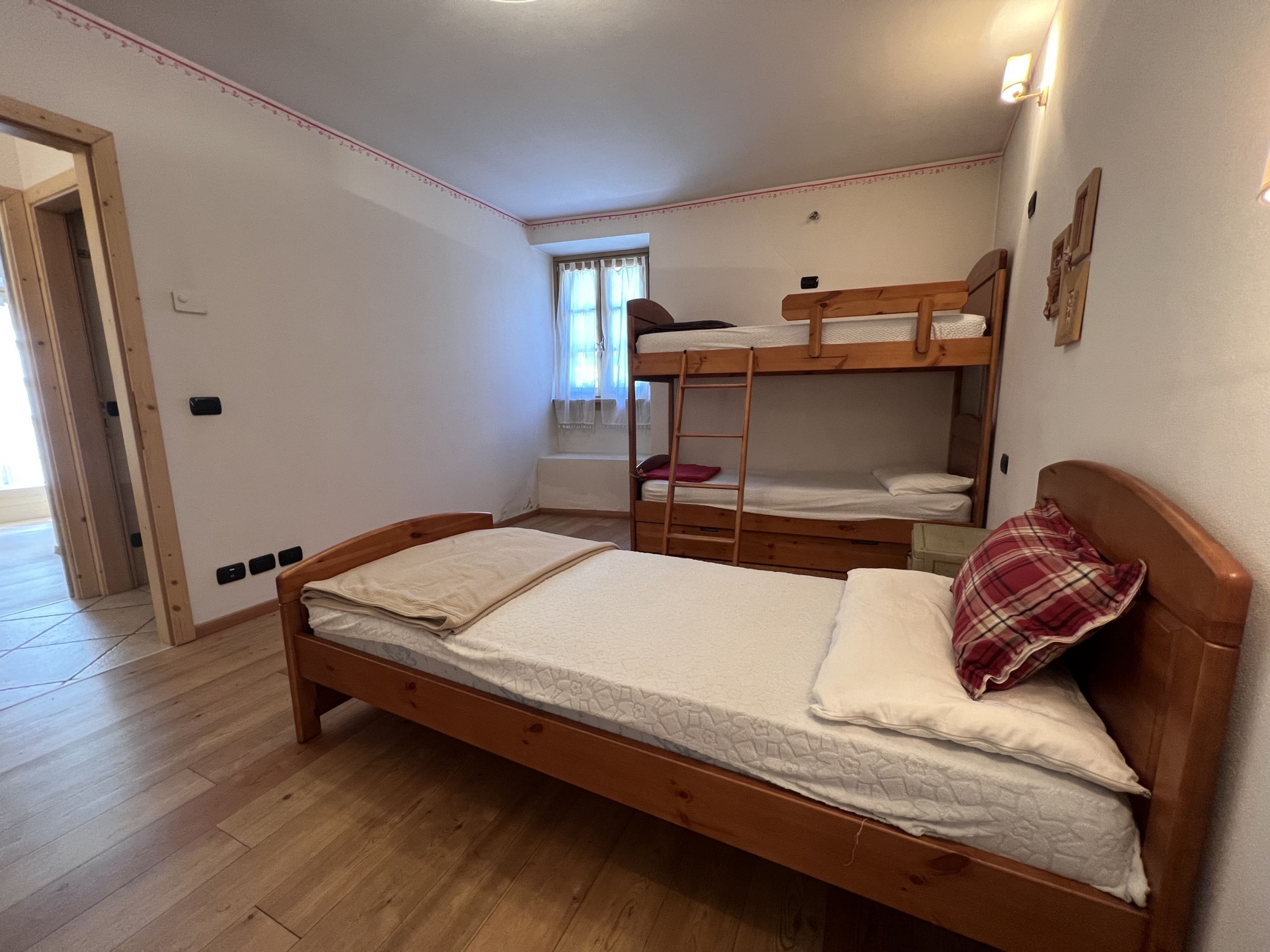 A bedroom with a single bed, a bunk bed, wooden flooring, a window with curtains, and a door.