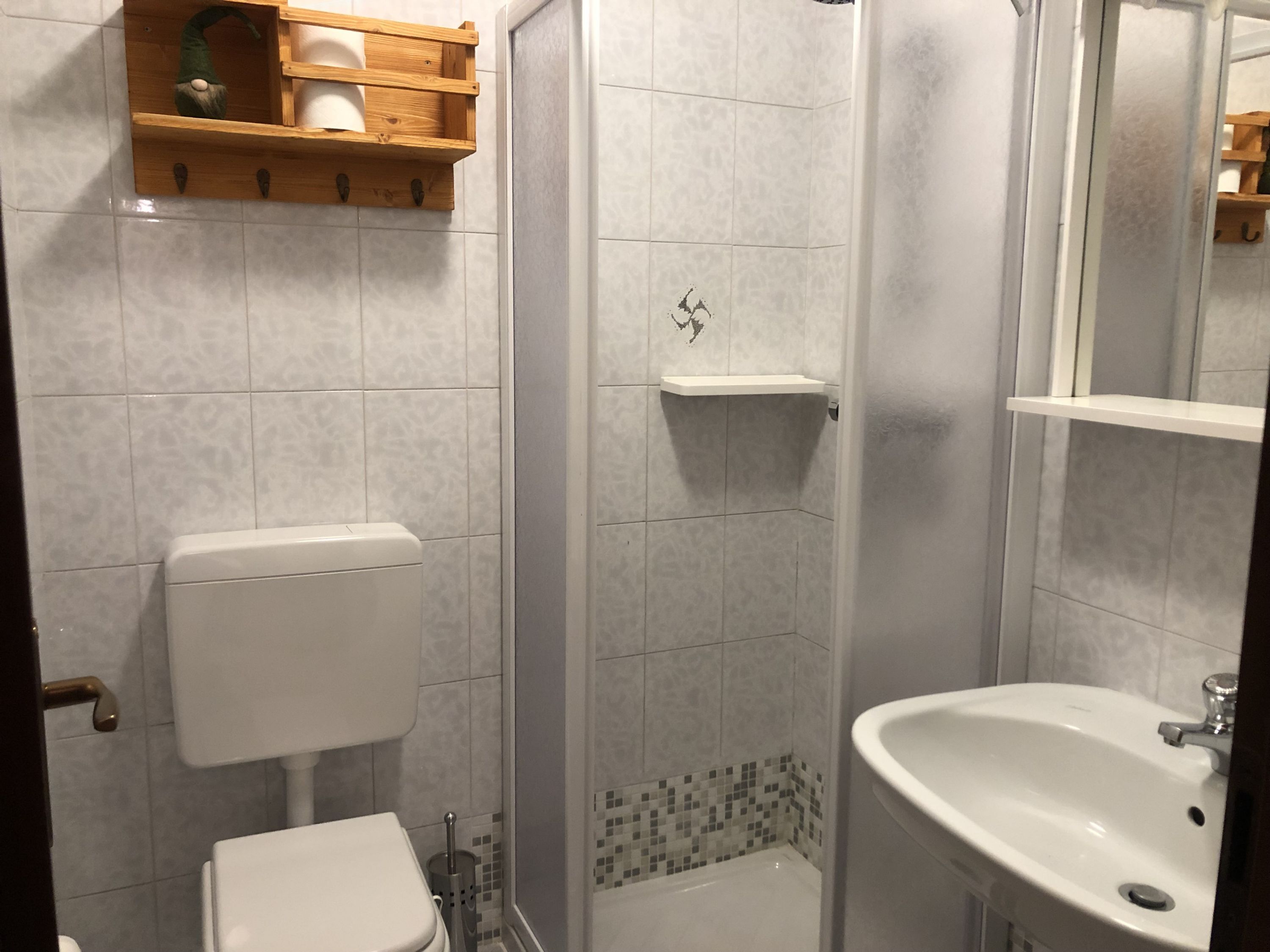 A bathroom with a toilet, sink, shower, and a shelf above the toilet.