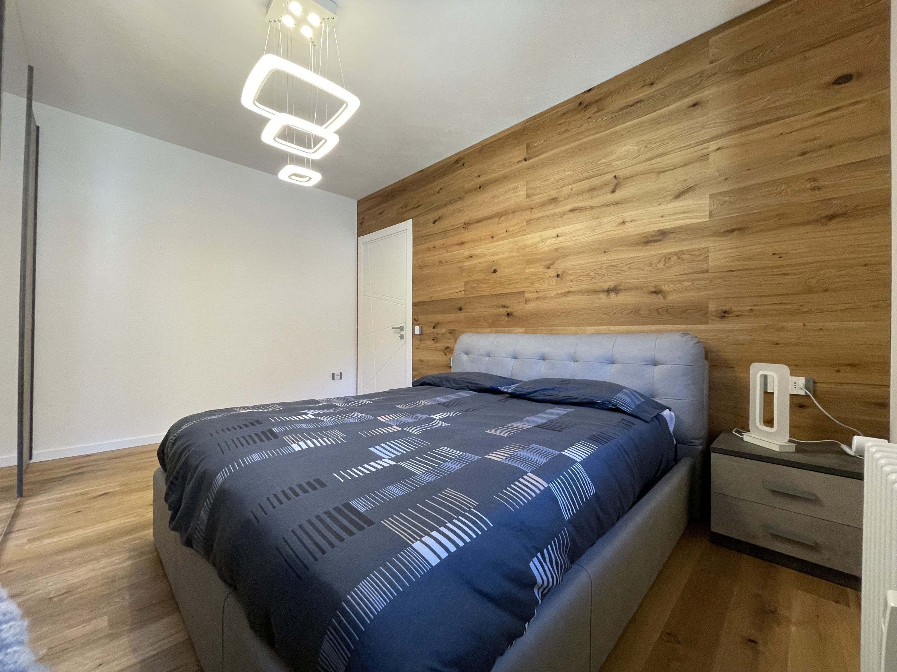 A modern bedroom with a wooden accent wall, a tufted headboard, a patterned bedspread, a bedside table with a lamp, and a power outlet.