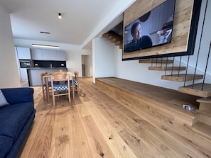 A modern living room with a wooden floor, a dining area with a table and chairs, a TV mounted on a wooden wall, and a kitchen in the background.