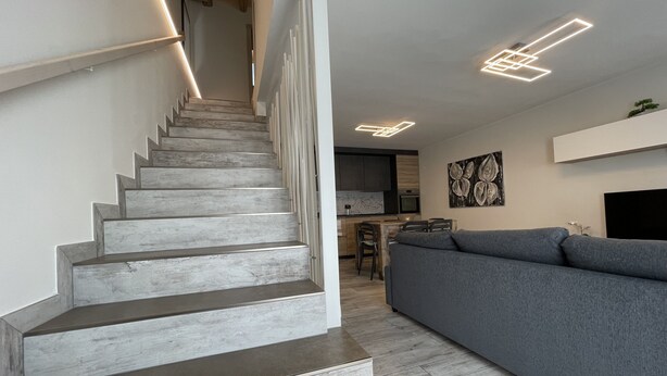 Un escalier moderne mène à un espace cuisine agrémenté d’un canapé gris et d’une table à manger.