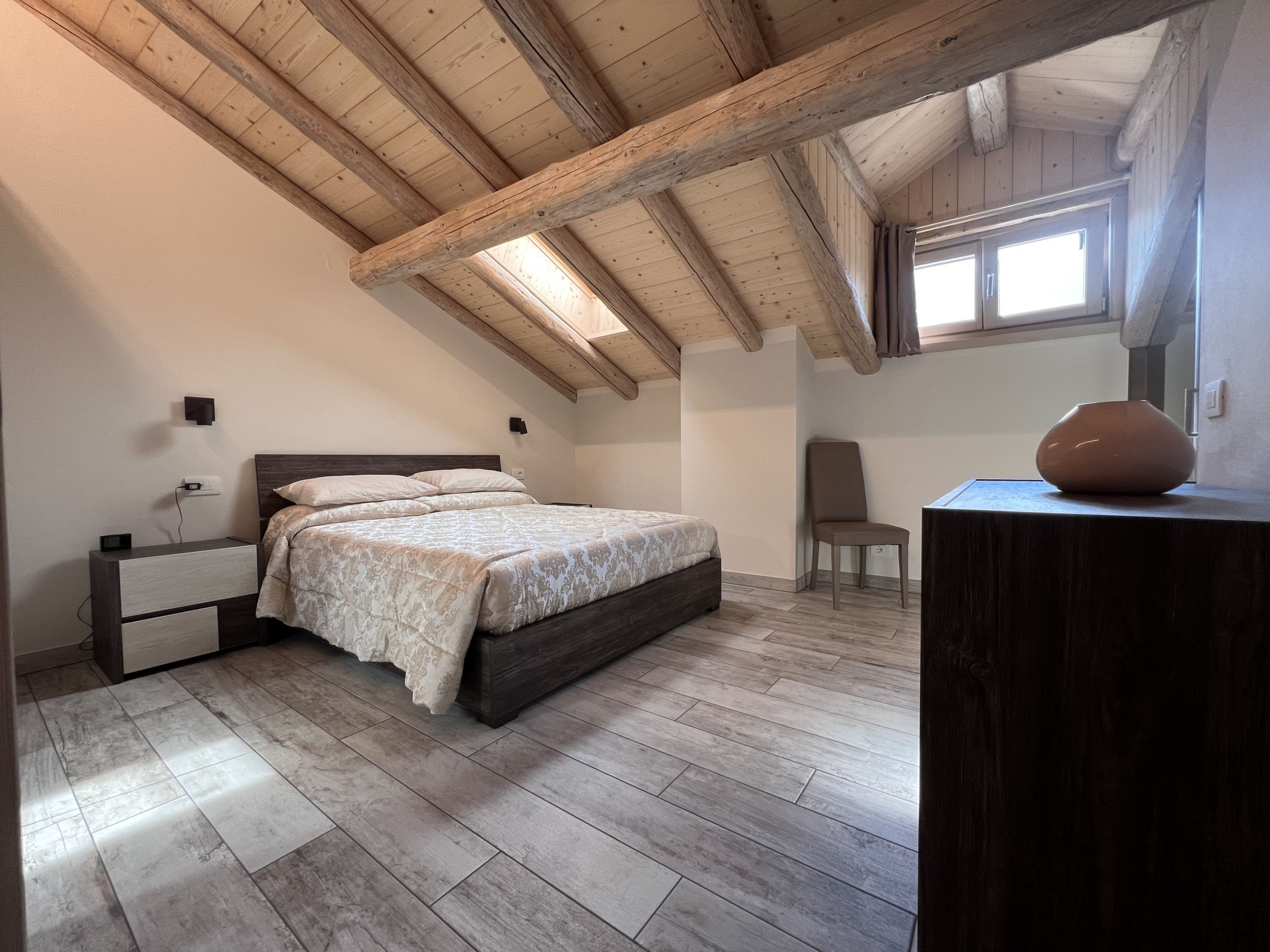 A bedroom with a wooden ceiling, a bed with patterned bedding, a bedside table, a chair, and a vase on a dresser.