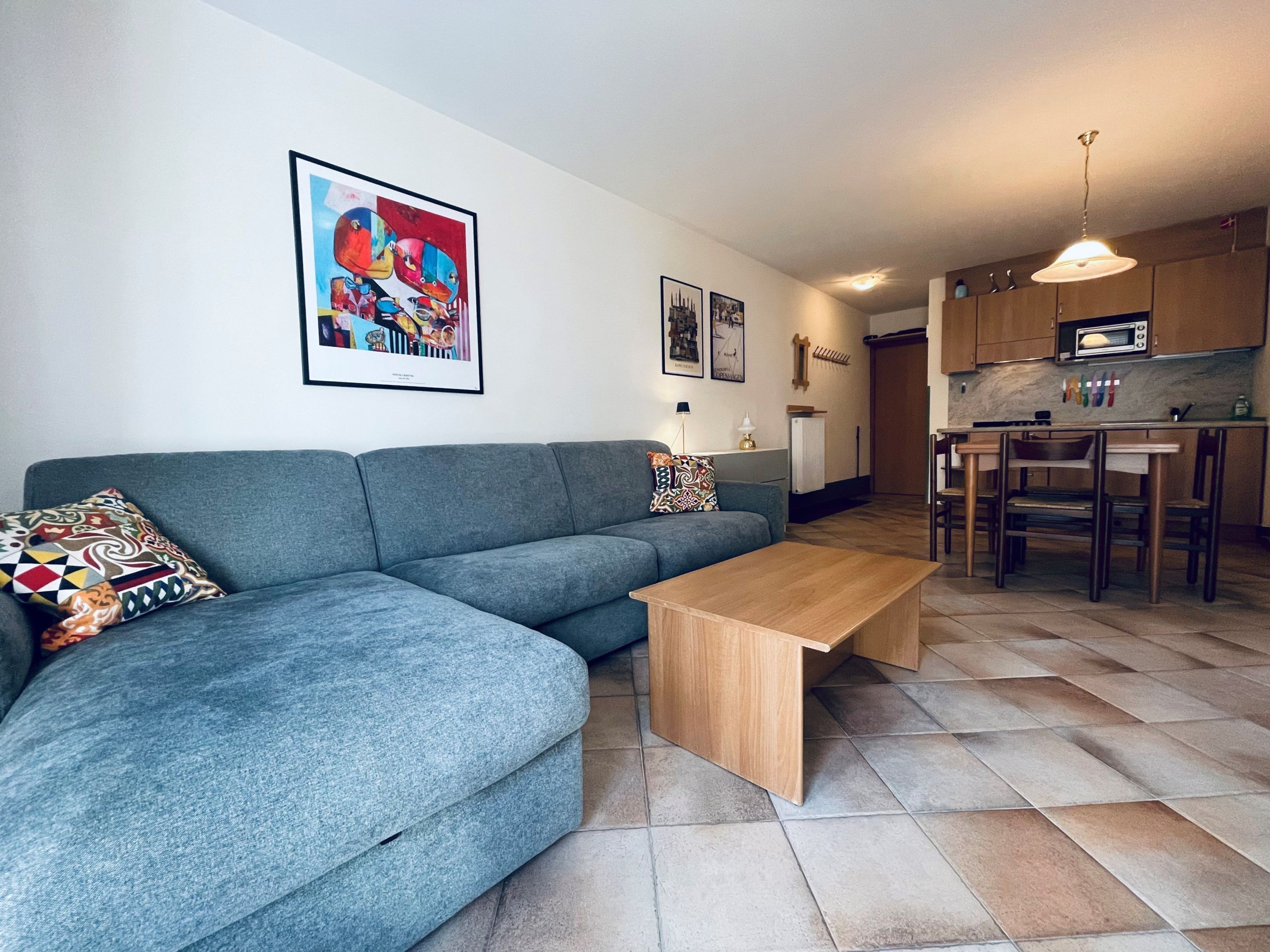 A modern living room with a grey sofa, a wooden coffee table, and a kitchen area with wooden cabinets and a microwave.