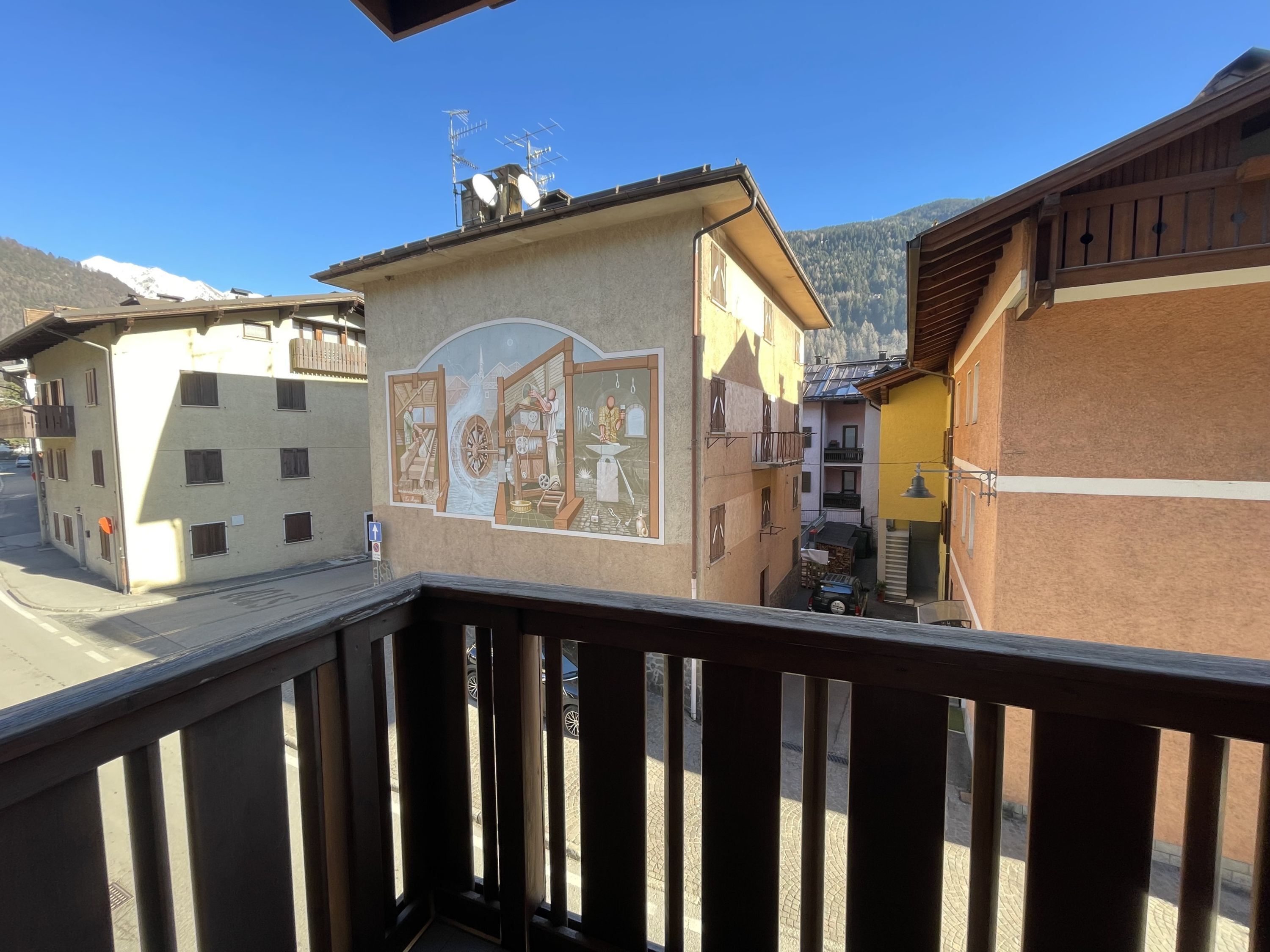 A building with a mural depicting traditional scenes, a balcony with a wooden railing, and a mountain backdrop.