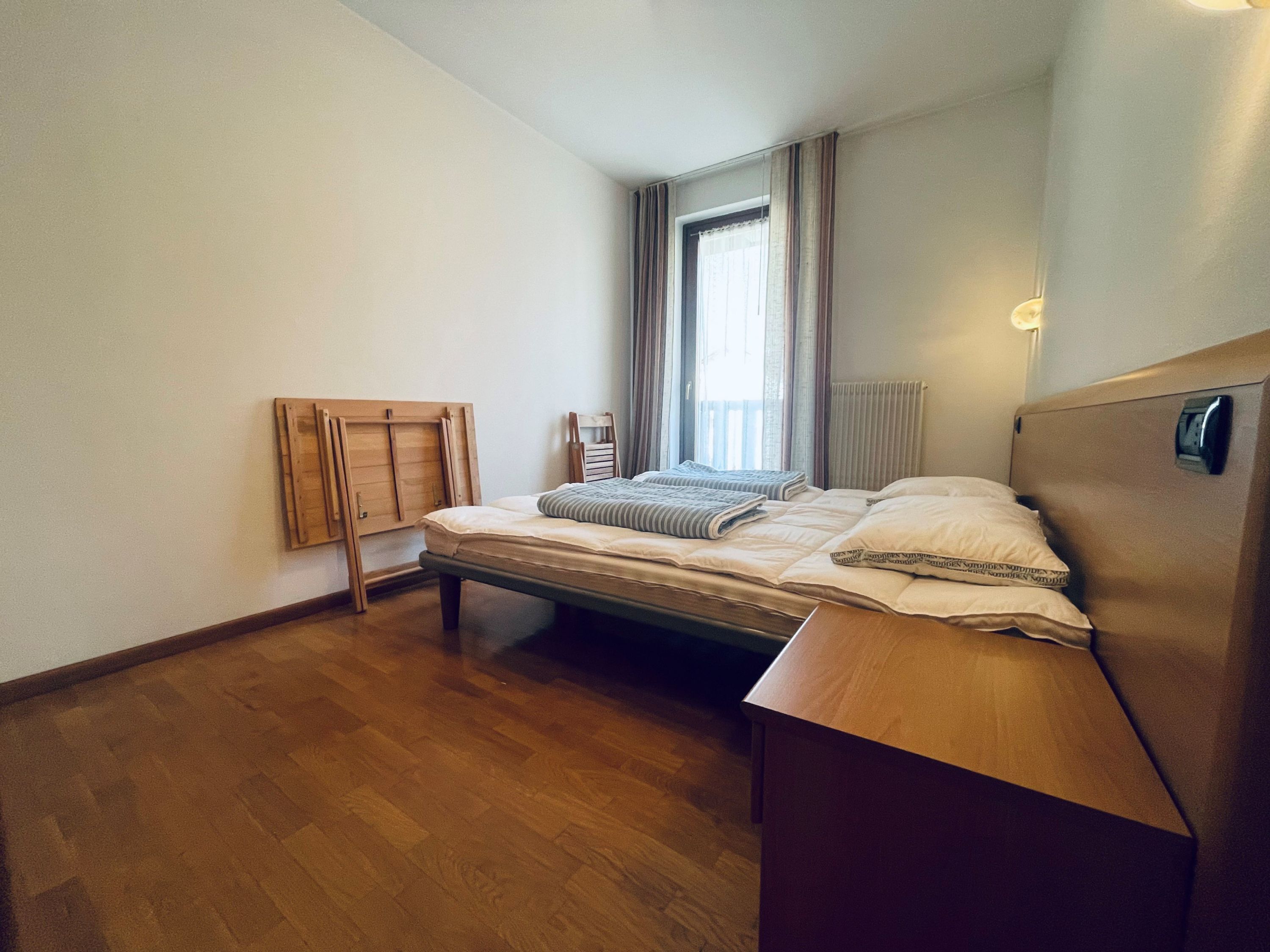 Una camera da letto con un letto di legno, una piccola sedia di legno, una finestra con tende e un termosifone.