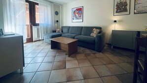 Un soggiorno con un divano grigio, un tavolino in legno e un pavimento piastrellato. Si notano due quadri appesi alla parete, una lampada su un tavolino laterale e una porta che dà su un'area esterna.