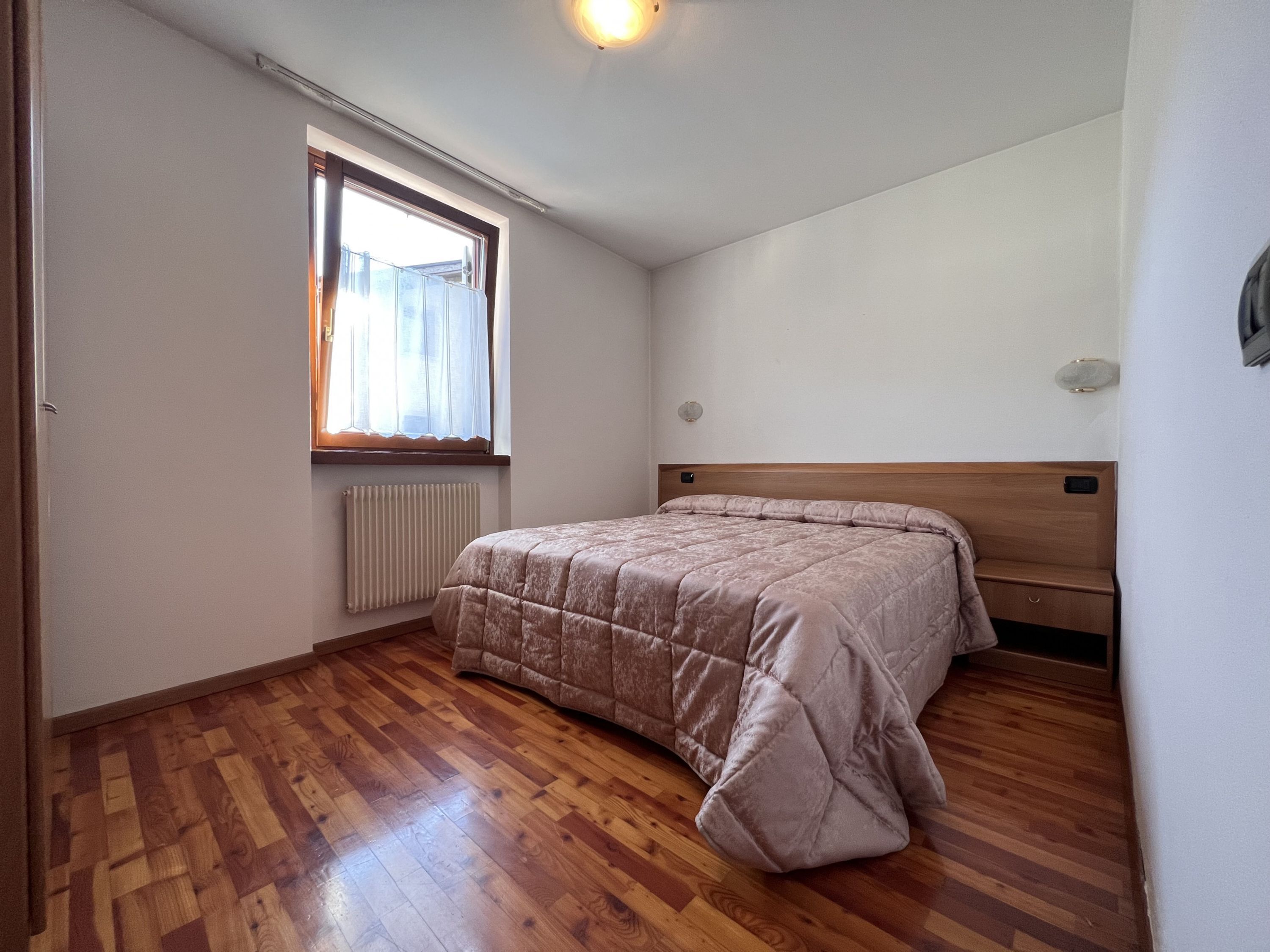 Une chambre avec un plancher en bois, un lit avec une tête de lit, une fenêtre avec des rideaux et un radiateur.
