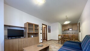 A living room with a sofa, a TV, wooden cabinets, a dining table, and a kitchen area with a microwave.
