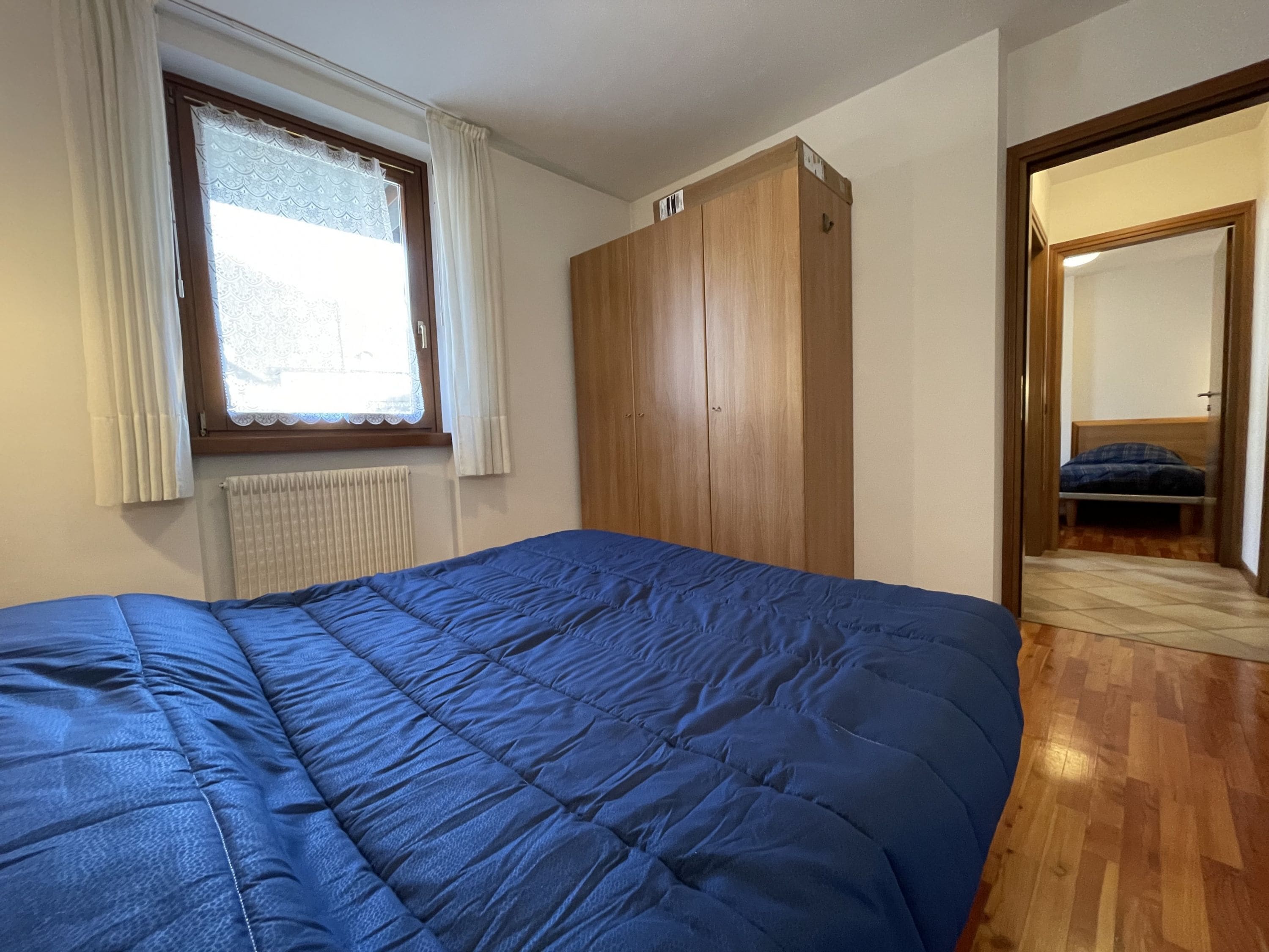 A bedroom with a large bed, a wardrobe, a radiator, and a window with curtains.