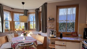 Een gezellige kamer met een bed, een eettafel met stoelen, een keuken met een fornuis en grote ramen met gordijnen.