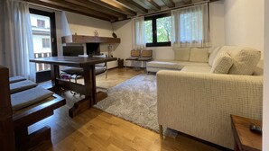 A cozy living room with a wooden ceiling, a fireplace, a sofa, a dining table, and a view of the outside through large windows. - 033 Trilocale Carisolo (Carisolo)