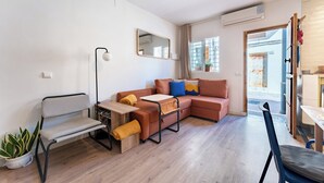 A compact living space with a sofa, a chair, a small table, and a floor lamp. There is a potted plant and a window with curtains. - Sunset House (Málaga)