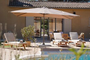 Een zwembadterras met ligbedden en een grote parasol, een plant in een pot en een gebouw met pannendak op de achtergrond.