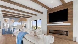 Een moderne woonkamer met een grote televisie, houten balken, een open haard en een keuken met witte kasten en een bar.