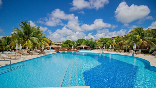 Una piscina grande y cristalina de color azul, rodeada de sillas de descanso y palmeras, bajo un cielo despejado.