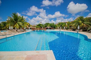 Una piscina grande y cristalina de color azul, rodeada de sillas de descanso y palmeras, bajo un cielo despejado.