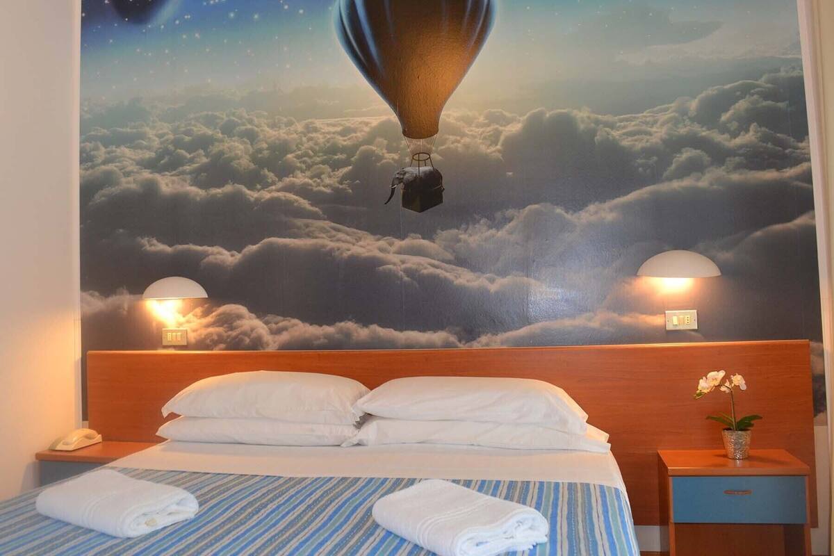 A hotel room with a bed, two bedside lamps, a nightstand, and a wall mural of clouds and a hot air balloon.
