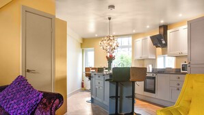 Un salon moderne avec un canapé, une table basse et un fauteuil jaune vif. On aperçoit à l’arrière-plan une cuisine dotée d’un îlot central et un coin repas avec une chaise et un vase de fleurs.